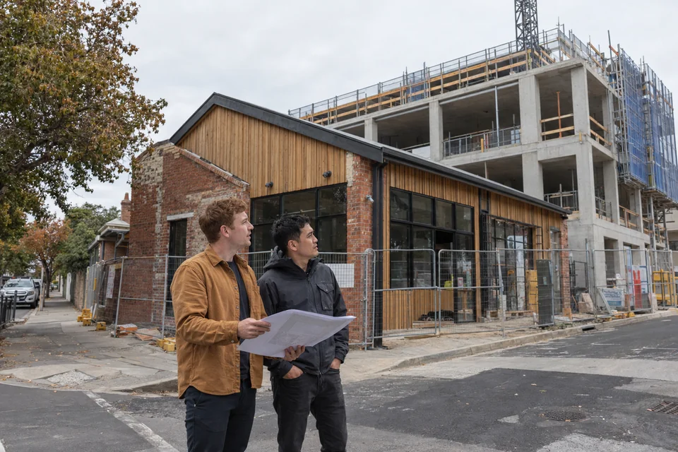 Illustrative scene: two building professionals compare a section drawing with a low mixed-use building beside a larger structure under construction.